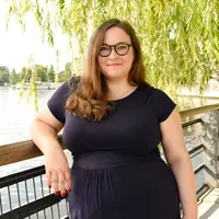 Portrait von Kristina Müller, die ein marineblaues Kleid trägt, steht an einem hölzernen Geländer am Wasser, über dem grüne Weidenzweige hängen. Sie lächelt in die Kamera.