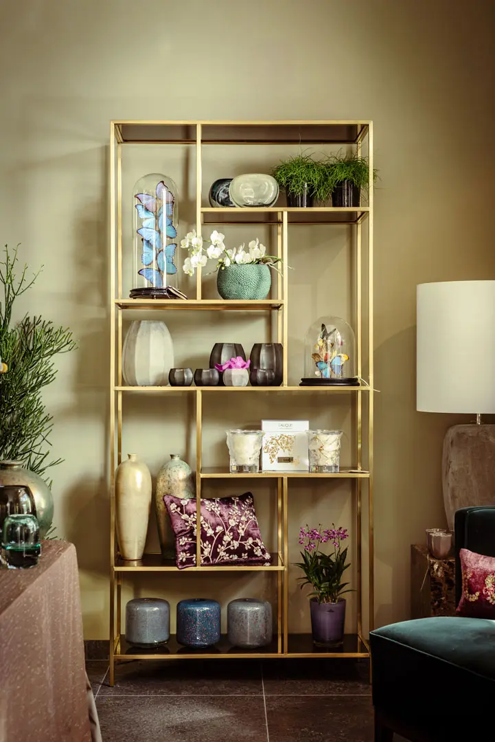 Ein goldenes Bücherregal aus Metall in einem Showroom enthält dekorative Gegenstände wie Vasen, Topfpflanzen, Kerzen, Glaskuppeln, Bücher, Blumenkissen und kleine Ornamente, die in einer gemütlichen, gut beleuchteten Wohnzimmerecke ordentlich angeordnet sind.