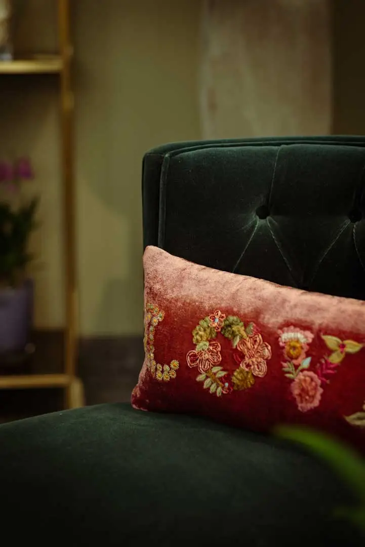 Ein dunkelgrüner Samtstuhl im Showroom von Blumen Bahlmann mit getufteter Rückenlehne und einem rosafarbenen Samtkissen, das mit bunten Blumenmustern bestickt ist, auf denen LOVE steht. Im Hintergrund sind eine verschwommene Pflanze und ein Regal zu sehen.