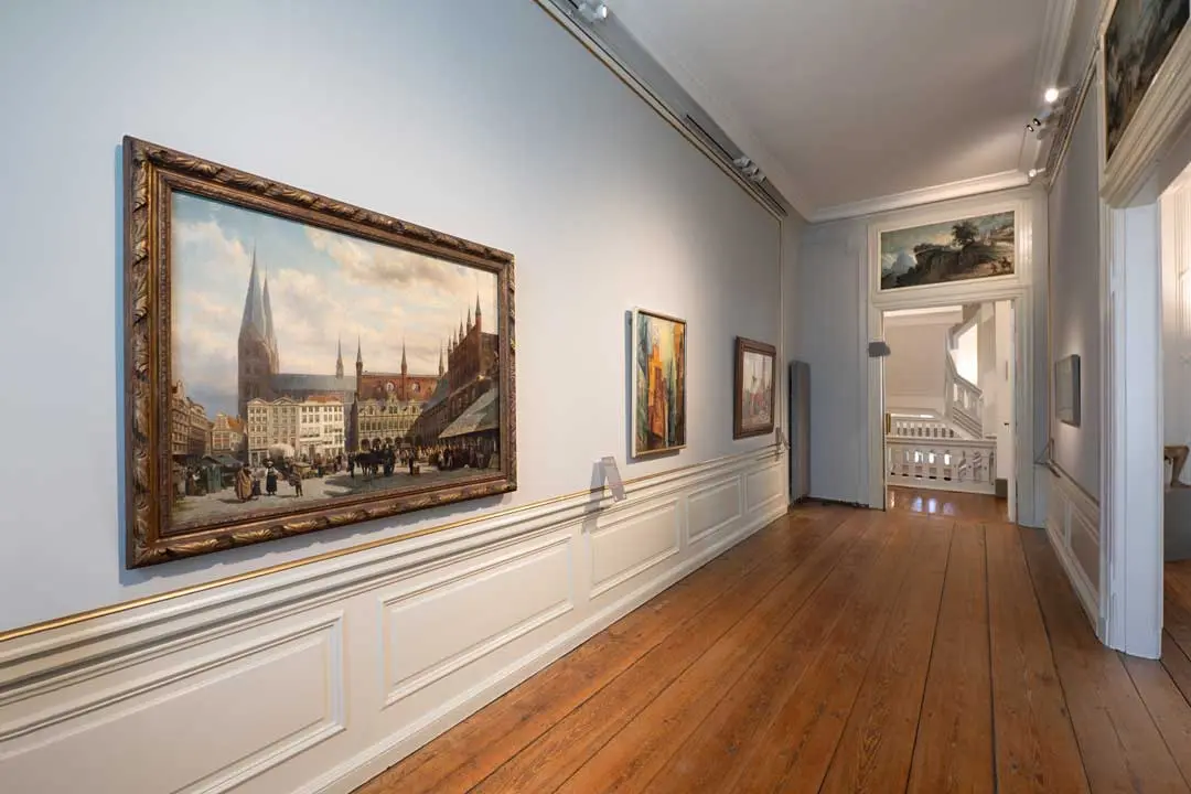 Eine Museumsfotografie zeigt einen Ausstellungsraum in einem Kunstmuseum mit Holzböden, weiß getäfelten Wänden und gerahmten Gemälden, darunter eine große Stadtlandschaft mit einer Kathedrale, die an der linken Wand hängt. Helles natürliches Licht fällt durch die Türöffnungen ein.