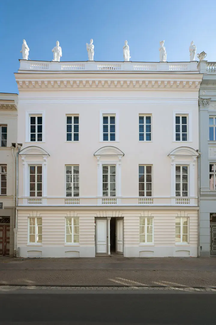 Fassade des Museums Behnhaus-Drägerhaus in Lübcke. Neoklassizistisches dreistöckiges Gebäude mit hellbeigen Wänden, gewölbten und rechteckigen Fenstern und weißen Statuen auf dem Dach vor einem klaren blauen Himmel.