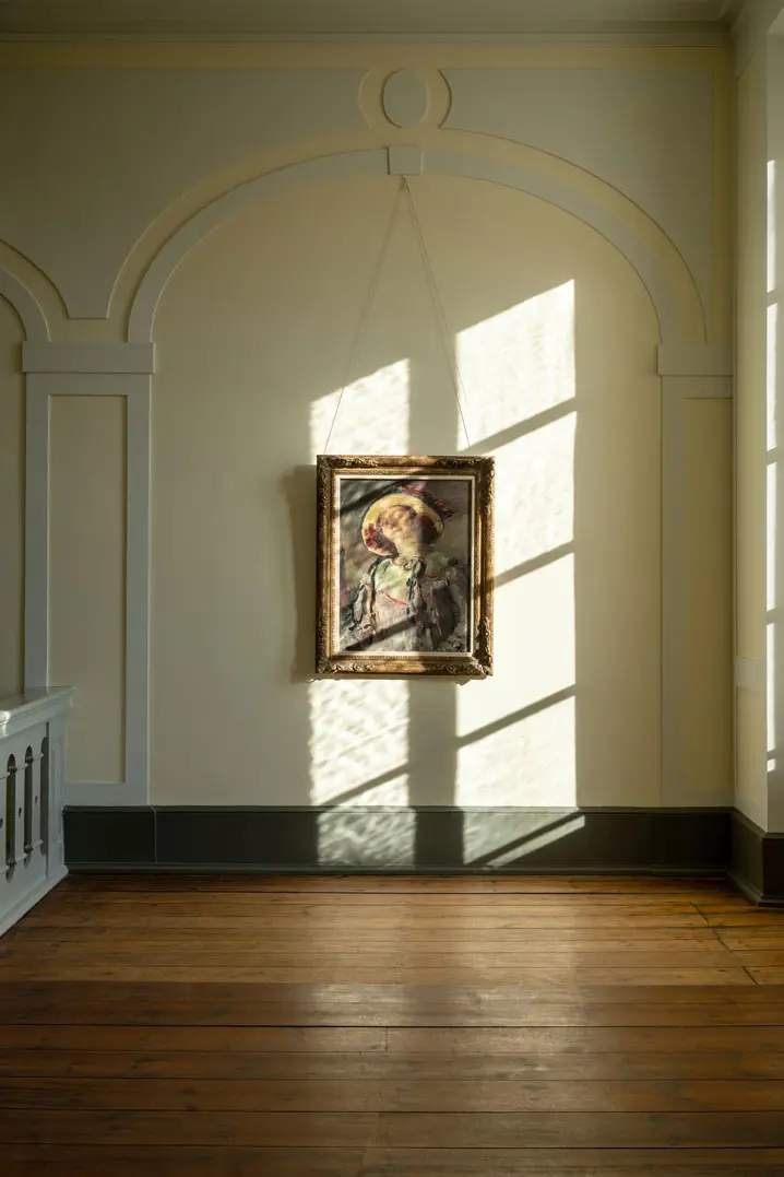 Ein gerahmtes Gemälde von Lovis Corinth hängt an einer cremefarbenen Wand im Museum Behnhaus-Drägerhaus, die von dem durch ein Fenster einfallenden Sonnenlicht beleuchtet wird, das Schatten und Lichtmuster wirft. Der Raum hat einen Holzboden und dekorative Wandleisten.