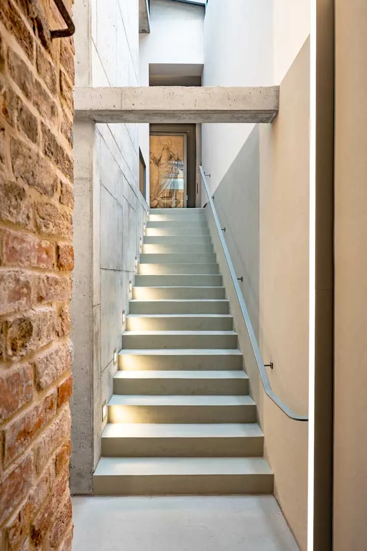 Eine moderne Treppe mit weißen Stufen führt zwischen Sichtziegeln und glatten Betonwänden nach oben. Sie ist mit einem eleganten Handlauf und einer eingebauten Stufenbeleuchtung ausgestattet und endet an einer Tür, hinter der ein Kunstwerk zu sehen ist.