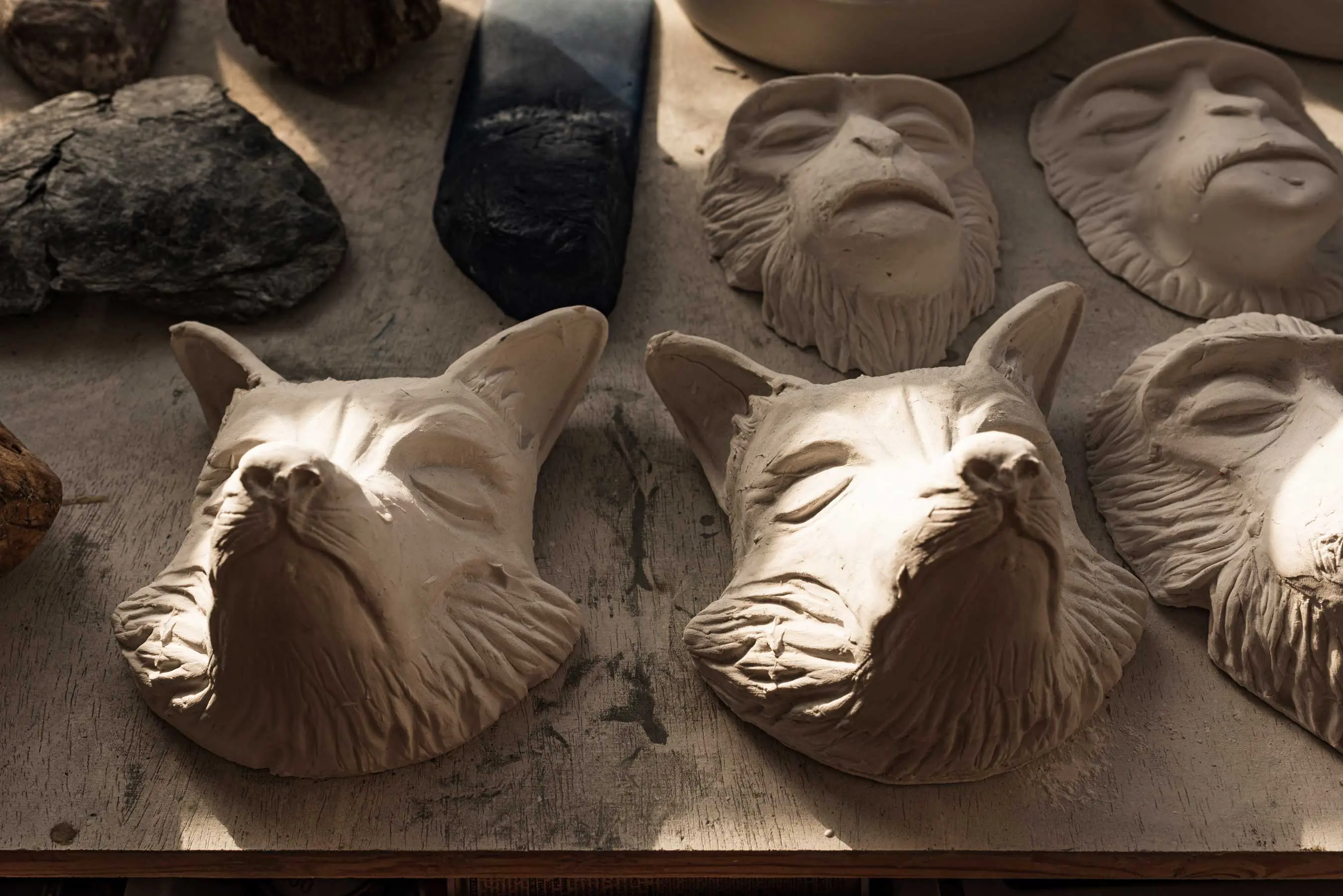 Auf einer hölzernen Fläche sind mehrere geschnitzte Tiermasken, darunter Füchse und Affen, aus einem hellen Material ausgestellt, auf die das Sonnenlicht Schatten wirft.