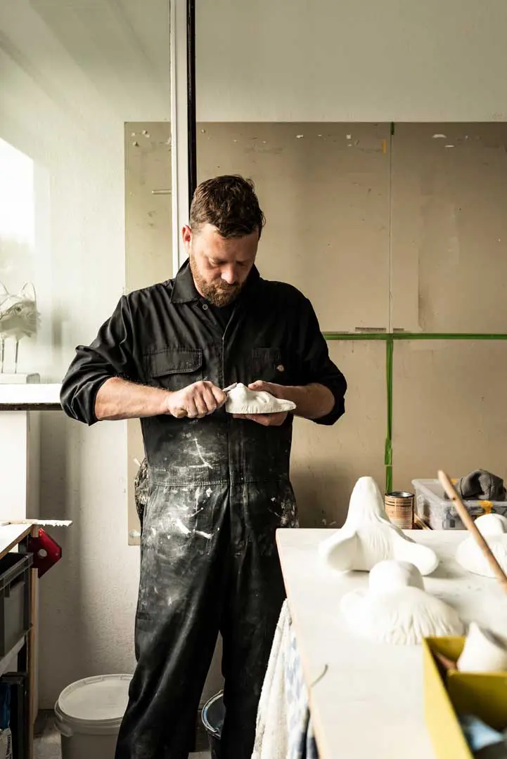 Der Künstler Björn Holzweg in einem schwarzen Overall arbeitet konzentriert an einer weißen Skulptur in einem hellen, unordentlichen Atelier, umgeben von Werkzeugen und unvollendeten Kunstwerken auf Tischen.
