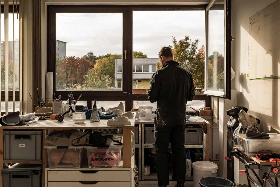 Björn Holzweg steht an einer Werkbank in einem gut beleuchteten Raum mit großen Fenstern und blickt nach draußen. Das Künstleratelier ist mit verschiedenen Werkzeugen, Behältern und Kunstzubehör vollgestopft, und durch das offene Fenster sind Bäume zu sehen.