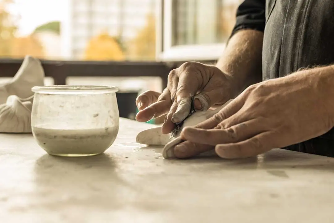 Nahaufnahme der Hände einer Person, die auf einem Tisch Ton formen, mit einem Glasgefäß mit Flüssigkeit in der Nähe. Der Hintergrund ist leicht unscharf und suggeriert einen Arbeitsbereich in einem Raum mit natürlichem Licht und Bäumen draußen.