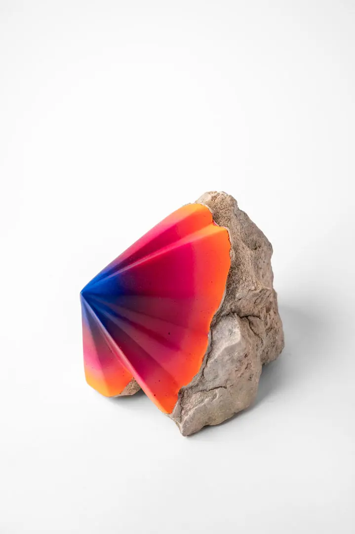 Auf einem rauen, hellen Felsen befindet sich ein buntes, fächerförmiges Objekt mit einem Farbverlauf von Blau über Rosa bis Orange, das einen auffälligen Kontrast zu einem schlichten weißen Hintergrund bildet.