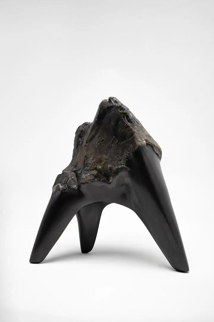 Eine Skulptur von Björn Holzweg mit einer rauen, felsigen Oberseite geht in drei glatte, schwarze, spitze Beine über, die an eine Mischung aus Naturstein und abstrakter, moderner Skulptur vor einem schlichten weißen Hintergrund erinnern.