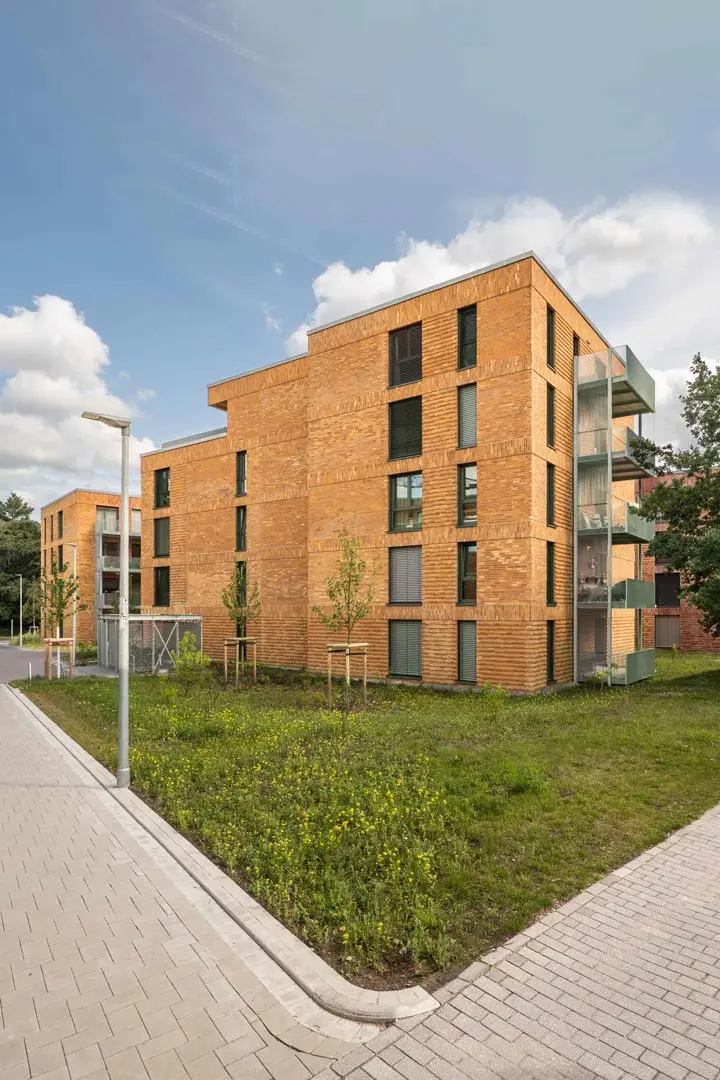 Architekturfotografie moderner Flachbauten mit Klinkerfassaden und großen Fenstern stehen neben einem gepflasterten Gehweg, einer Rasenfläche und jungen Bäumen unter einem teilweise bewölkten Himmel. Realisiert von Blu by August Prien.