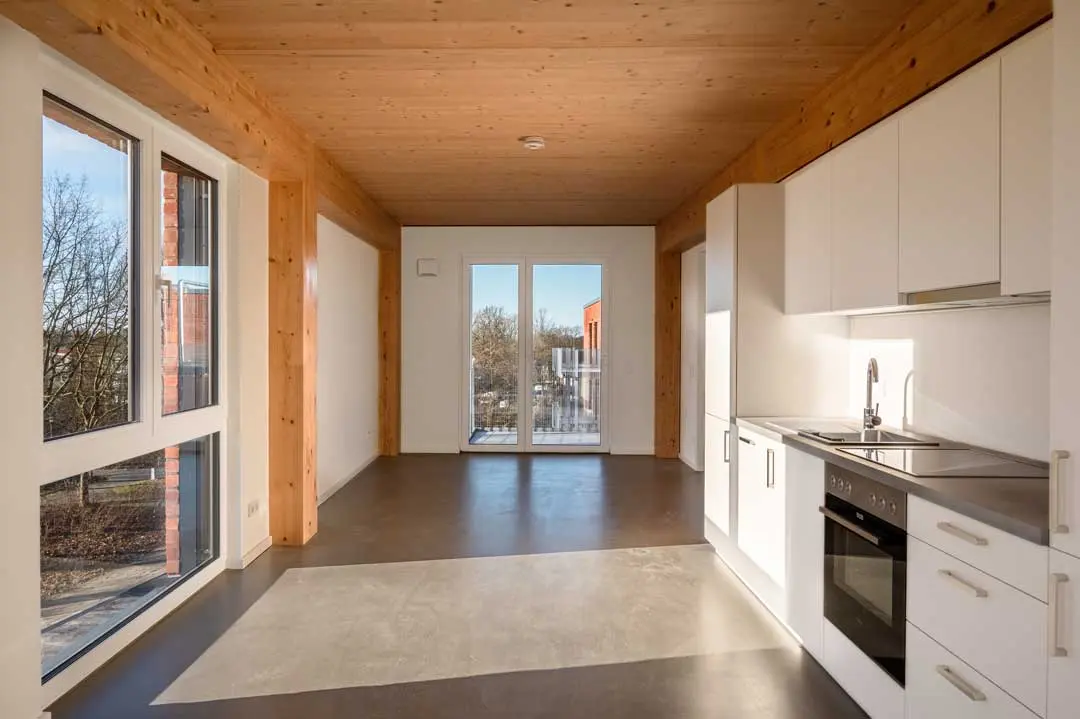 Ein modernes, unmöbliertes Zimmer mit großen Fenstern, einer Holzdecke, weißen Küchenschränken, einem eingebauten Ofen und Glastüren, die auf einen Balkon führen. Natürliches Licht füllt den hellen, minimalistischen Raum.