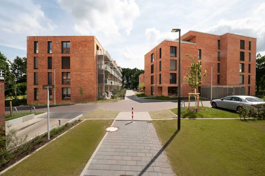 Moderne Wohnhäuser mit roten Backsteinfassaden stehen auf beiden Seiten eines gepflasterten Weges, umgeben von grünem Rasen, jungen Bäumen und einer Straßenlaterne unter einem teilweise bewölkten Himmel.