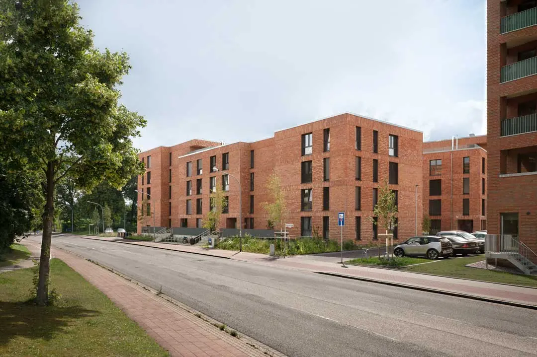Ein modernes Wohngebäude, errichtet von Blu by August Prien mit Backsteinfassade und mehreren Stockwerken liegt an einer ruhigen Straße mit geparkten Autos, Bäumen und einem breiten Gehweg unter einem teilweise bewölkten Himmel.