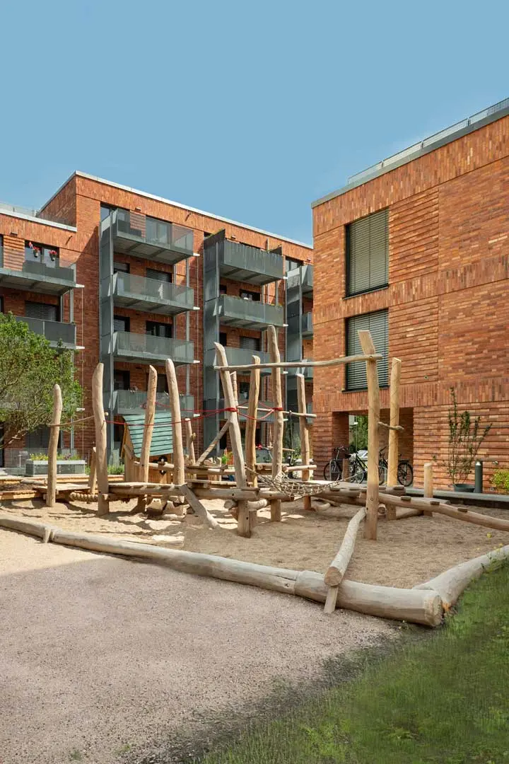 Ein moderner Spielplatz mit hölzernen Klettergerüsten liegt auf einer sandigen Fläche zwischen zwei Backsteinwohnhäusern unter einem strahlend blauen Himmel.