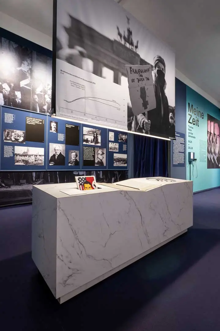 Eine moderne Museumsausstellung der Ausstellung über Thomas Mann mit einem Marmortisch, historischen Fotos und blauen Informationstafeln an den Wänden. Dieses Ausstellungsfoto der Ausstellung über Thomas Mann zeigt einen Marmortisch, historischen Fotos und blauen Informationstafeln an den Wänden. Darüber hängt ein großes Schwarz-Weiß-Foto, auf dem ein Demonstrant ein Schild hält.
