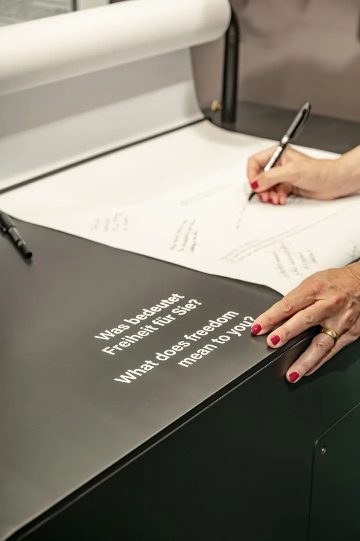 Eine Person mit rotem Nagellack schreibt auf ein großes Blatt Papier. Im Vordergrund steht auf einer schwarzen Fläche der Text "Was bedeutet Freiheit für Sie?" in Englisch und Deutsch.