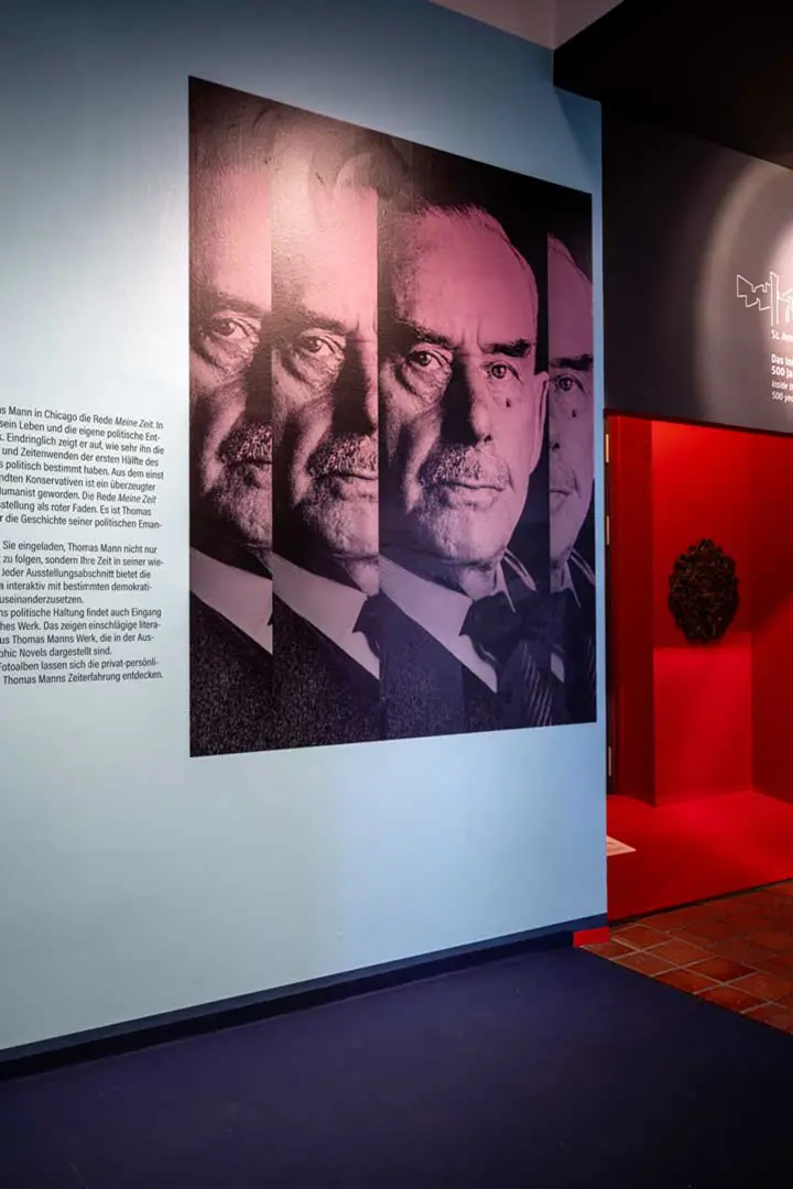 Diese Ausstellungsfotografie zeigt ein Wanddisplay auf dem ein großes, künstlerisches Porträt von Thomas Manns, das in überlappenden, allmählich verblassenden Schichten dargestellt ist. Neben dem Bild ist ein Textblock zu sehen. Im Hintergrund sind ein roter Flur und ein gefliester Boden zu sehen.