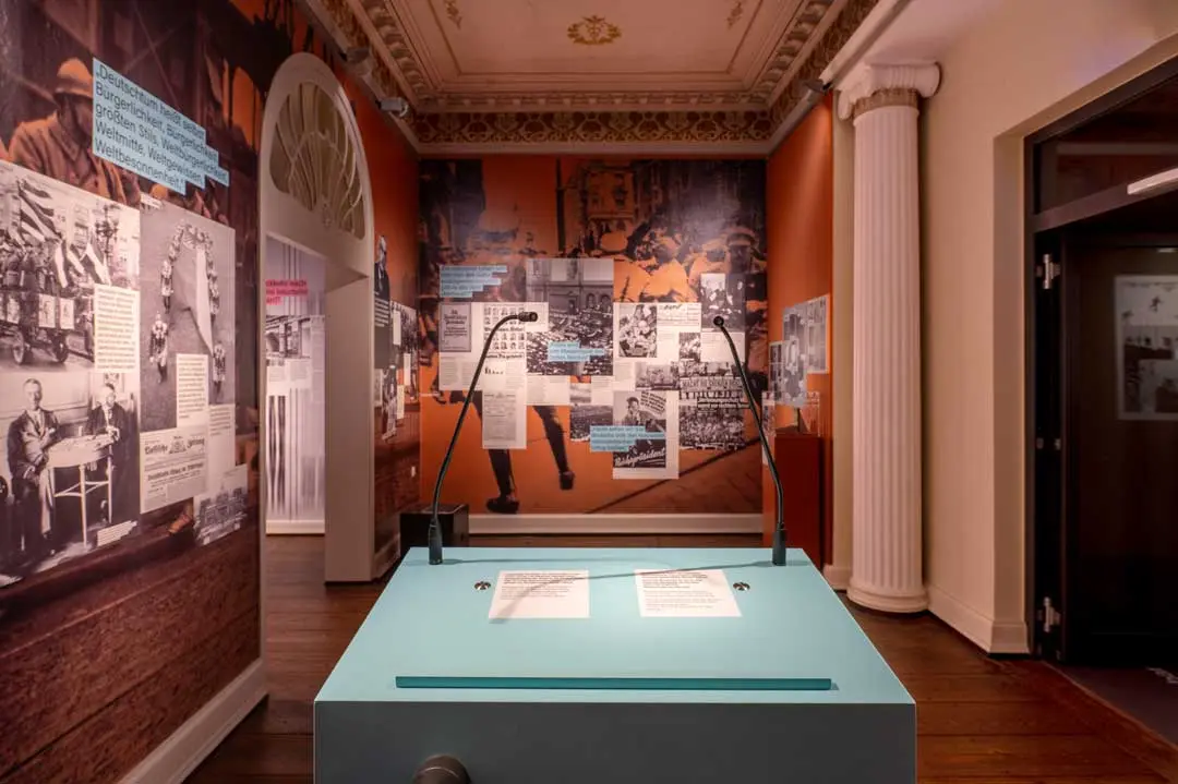 Eine Ausstellungsfotografie zeigt einen Ausstellungsraum der Ausstellung "150 Jahre Thomas Mann". Es sind historische Fotografien, Dokumente und blau hervorgehobene Texttafeln an den Wänden zu sehen mit einer zentralen blauen Vitrine und verzierten architektonischen Details wie Säulen und einer dekorativen Decke.