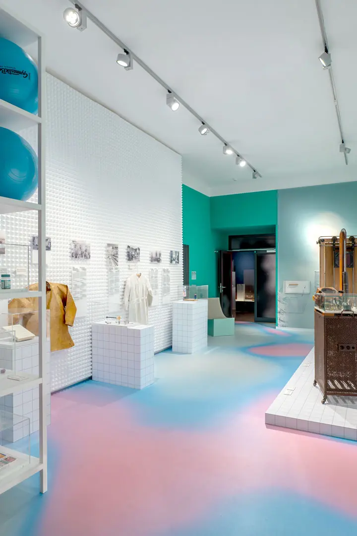 Foto einer Ausstellungsdokumentation zeigt einen Ausstellungsraum mit weiß gekachelten Auslagen, alter Kleidung, Fotos an den Wänden und wissenschaftlichen Instrumenten. Die Wände sind blaugrün, und der Boden hat einen Farbverlauf aus blauer und rosa Beleuchtung.