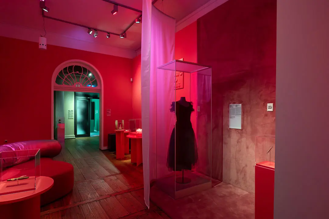 Ein Ausstellungsraum eines Museums mit roten Wänden und Beleuchtung, in dem ein schwarzes Kleid in einer Glasvitrine, andere Artefakte auf Tischen und ein geschwungenes Sofa in der Ecke ausgestellt sind. Im Hintergrund ist ein gewölbter Durchgang zu sehen.