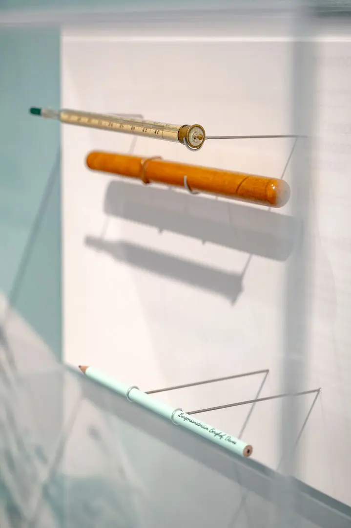Eine Vitrine mit zwei Schreibgeräten und einen Termometer, die jeweils horizontal mit durchsichtigen Halterungen aufgehängt sind; die Schreibgeräte variieren in Farbe und Design, wobei eines aus Holz und ein anderes in einem hellen Blau gehalten ist. Sie sind Teil einer Ausstellung des Buddenbrookhauses.