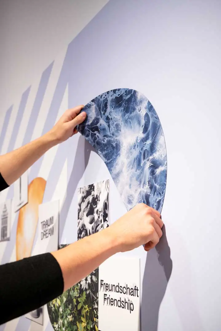 Die Hände einer Person befestigen einen geschwungenen, blau-weißen abstrakten Druck an einer Wand zwischen anderen Bildern und Texten, darunter die Worte "Freundschaft Friendship" und "TRAUM DREAM" zur Ausstellung Zauberberg.