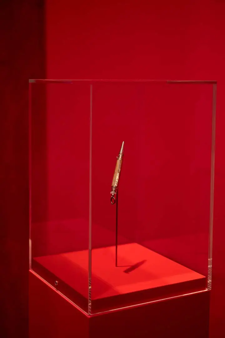 Ein Kugelschreiber, das aufrecht in einem durchsichtigen Glaskasten auf einem roten Sockel steht, umgeben von einem roten Hintergrund.