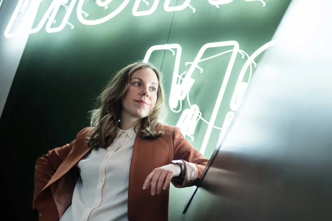 Eine Frau in einem braunen Blazer und weißem Hemd lehnt lässig für ein Business Foto an einer spiegelnden Fläche und schaut nachdenklich. Hinter ihr leuchtet ein Neonschild an einer dunklen Wand. Die Beleuchtung schafft eine moderne, entspannte Atmosphäre.