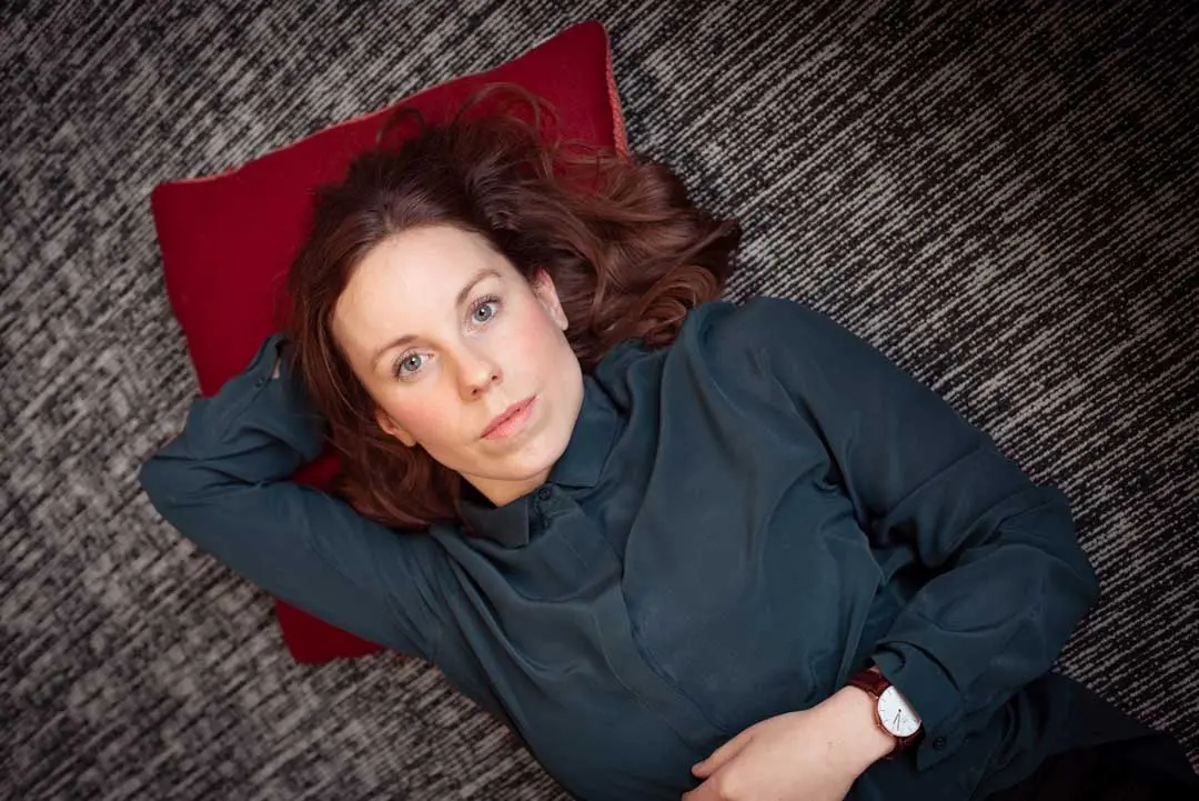Eine Frau mit braunen Haaren liegt auf einem roten Kissen auf einer grauen, strukturierten Oberfläche. Sie trägt ein dunkelgrünes Hemd und eine Uhr und schaut mit entspanntem Blick auf. Das Bild dient für ihre Businessfotografie und Personal Branding.