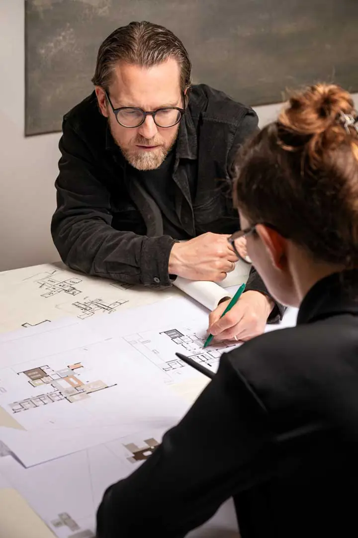 Ein Mann und eine Frau diskutieren über architektonische Grundrisse, die auf einem Tisch ausgebreitet sind. Der Mann beugt sich aufmerksam vor, während die Frau Notizen auf den Plänen macht. Beide scheinen sich auf die Diskussion zu konzentrieren und sich damit zu beschäftigen.