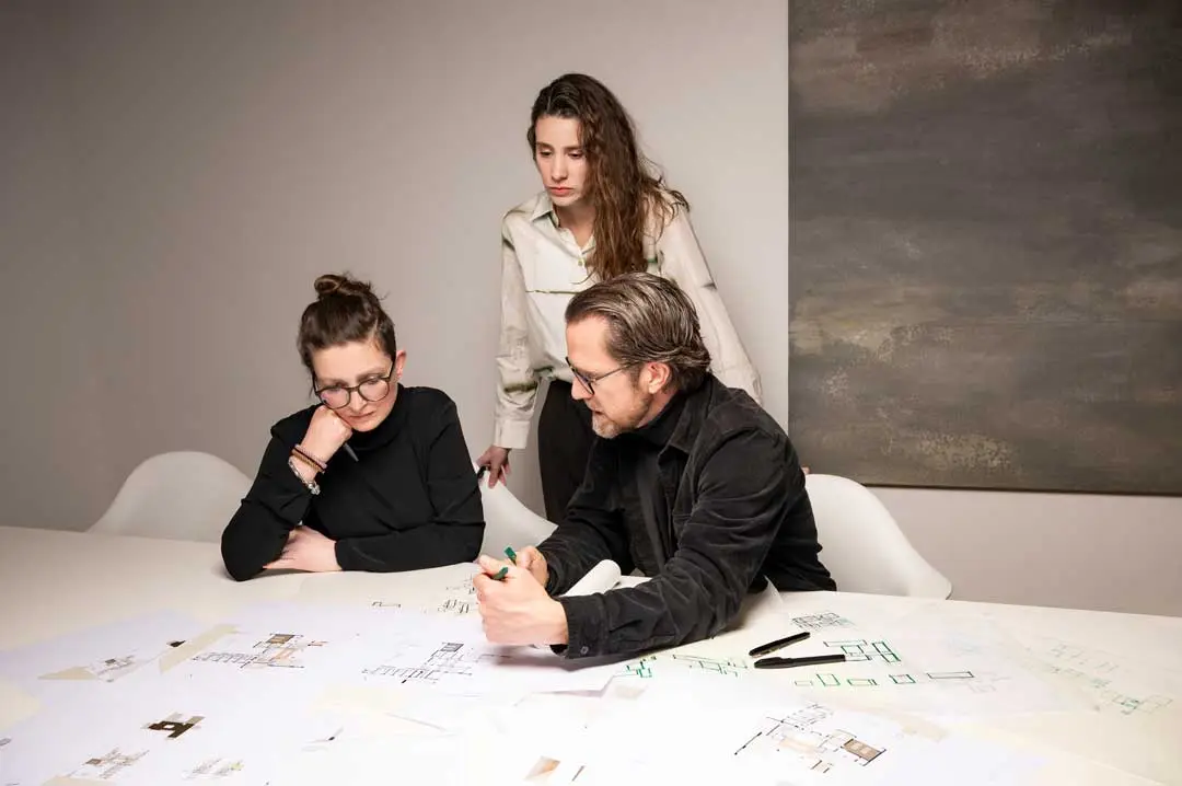 Drei Personen sitzen und stehen um einen weißen Tisch, der mit architektonischen Zeichnungen und Papieren bedeckt ist, und diskutieren über Pläne. Ein Mann erklärt einen Punkt, während zwei Frauen aufmerksam zuhören. Alle wirken konzentriert und engagiert.