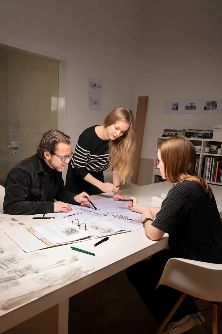 Für eine Corporate Fotografie sitzen und stehen drei Personen um einen Tisch herum, der mit Architekturzeichnungen und Stiften bedeckt ist, und besprechen ihre Arbeit in einer modernen Büroumgebung mit Bücherregalen und Wandfotos im Hintergrund.