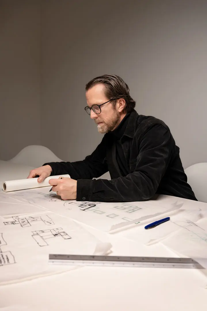 Der Architekt Daniel Volske sitzt für eine Corporate Fotografie an einem Tisch, der mit Architekturzeichnungen bedeckt ist, hält ein zusammengerolltes Papier in der Hand und prüft Baupläne. Ein Stift und ein Lineal liegen ebenfalls auf dem Tisch.