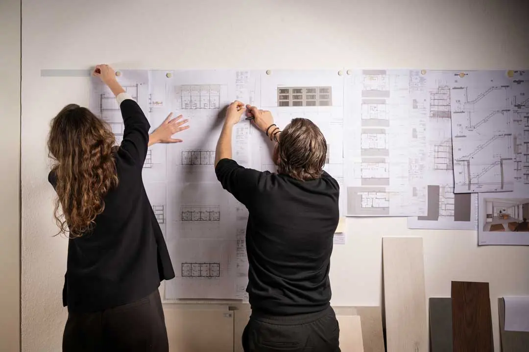 Das Team von Daniel Volske Architekten, die von hinten zu sehen sind, befestigen große architektonische Pläne und Zeichnungen an einer Wand und arbeiten zusammen, um die Dokumente zu ordnen und zu organisieren. In der Nähe sind verschiedene Papiere und Entwurfsmuster ausgebreitet.