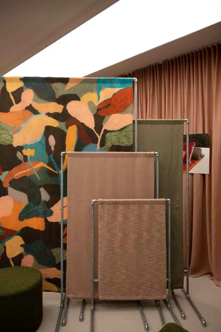 Drei Stoffbahnen in Erdtönen sind auf Metallständern vor einem großen, farbenfrohen, abstrakten Textilhintergrund in einem Raum mit beigen Vorhängen und gedämpfter Beleuchtung ausgestellt im Showroom Fischbacher 1819, Paris.