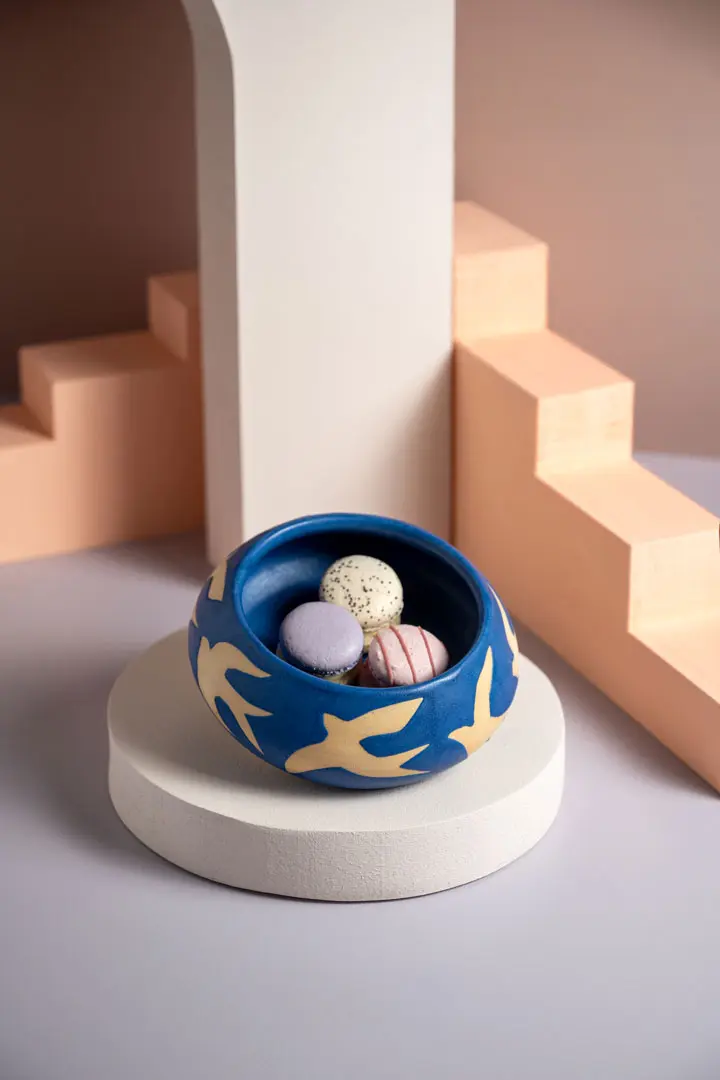 Eine blaue handgemachte Keramik Schale von Float Studio mit gelben Vogelmotiven hält vier bunte Macarons auf einem weißen Sockel. Pfirsichfarbene und weiße geometrische Treppen bilden den Hintergrund für eine moderne, künstlerische Stillleben-Fotografie-Szene.