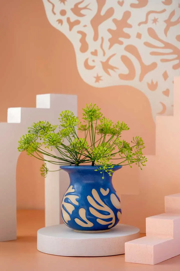 Eine blaue Vase von Float Studio Keramik mit cremefarbenen abstrakten Mustern hält grüne Blütenstängel. Diese Still-Life Fotografie zeigt geometrische Formen in Weiß und Rosa vor einem pfirsichfarbenen, gemusterten Hintergrund.