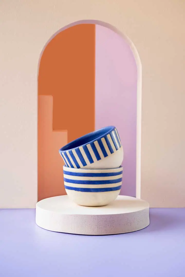 Zwei gestapelte Keramiktassen mit blauen Streifen und blauen vertikalen Linien stehen auf einem runden weißen Sockel vor einem bogenförmigen, pastellrosa, lila und orangefarbenen Hintergrund - eine heitere Float Studio Keramik Still-Life Fotografie Szene.