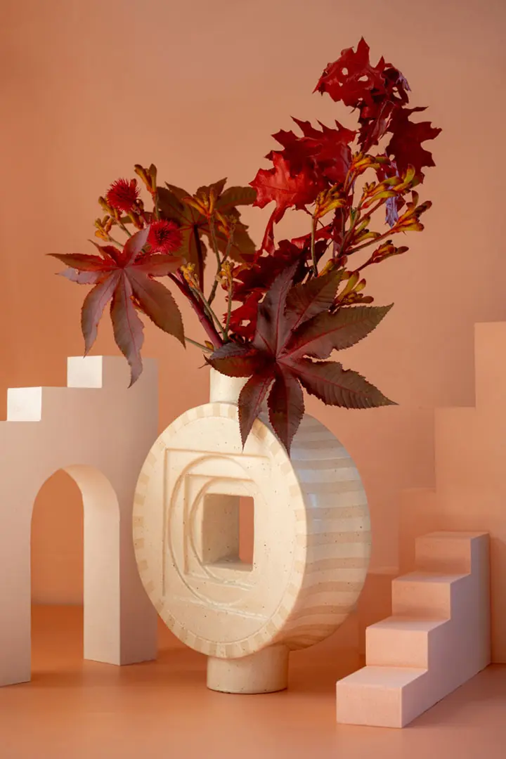 Eine moderne Keramik Vase von Float Atelier mit geometrischen Mustern hält rote Blätter und Blumen inmitten minimalistischer weißer architektonischer Formen, einschließlich Bögen und Treppen, vor einem warmen beigen Hintergrund in dieser eleganten Stilllebenfotografie.