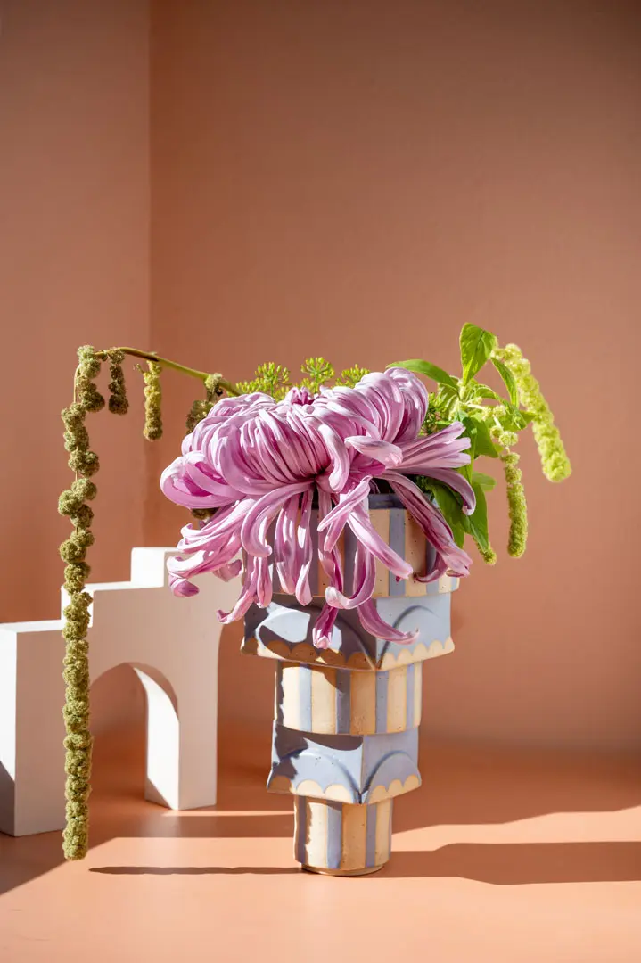 Elegante Still-Life Fotografie einer skulpturalen Vase von Float Atelier Hamburg mit blauen und cremefarbenen Akzenten enthält eine große, blassviolette Chrysantheme und einen grünen hängenden Amaranthus vor einem pfirsichfarbenen Hintergrund mit einem weißen geometrischen Bogen.