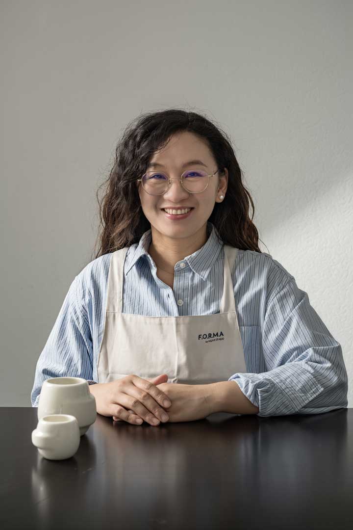 Die Designerin Hong Yi Zuh sitzt an einem Tisch und lächelt; sie trägt ein gestreiftes Hemd und eine beige Schürze. Vor ihr stehen drei weiße Keramiktassen auf dem dunklen Tisch.