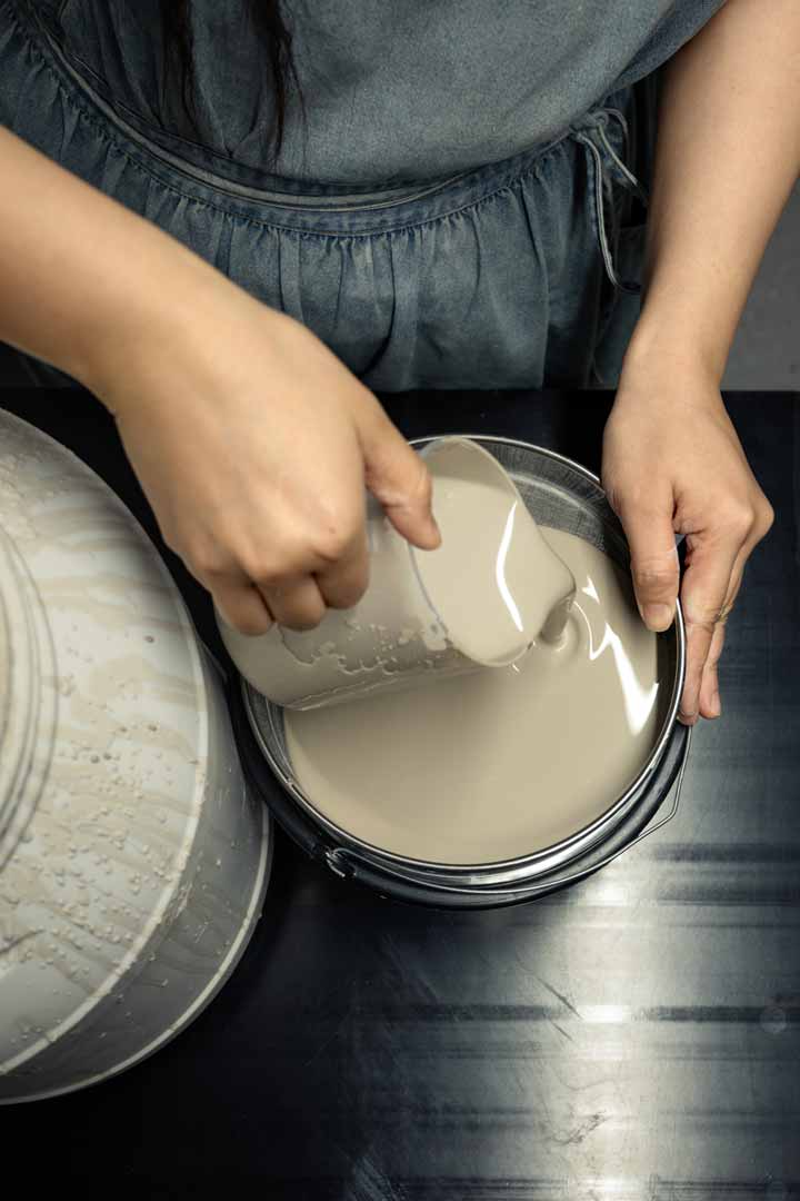 Eine Person in einer Jeans Schürze gießt Porzellanmasse aus einem Becher in einen Metalleimer auf einem dunklen Tisch.