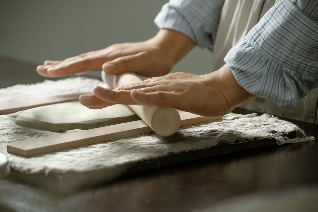 Die Designerin Hong Yi Zuh rollt Ton, der zwischen zwei Holzlatten auf einer mit einem Tuch bedeckten Fläche liegt, mit einem Nudelholz aus.