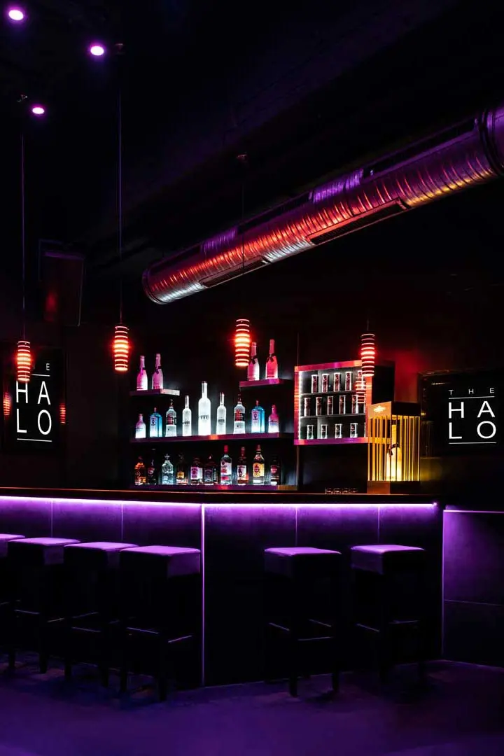 Eine moderne Bar des Clubs Halo mit leuchtenden lila- und rosafarbenen Lichtern, Flaschen in Regalen, hängenden Pendelleuchten, hohen Hockern am Bartresen und metallischen Luftkanälen an der Decke.