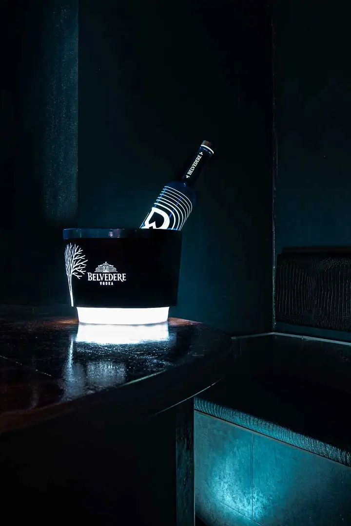 Eine Flasche Belvedere-Wodka kühlt in einem beleuchteten Eiskübel auf einem dunklen Holztisch, der in einem schummrigen, modernen Interieur mit dunklen Wänden und Sitzgelegenheiten steht.