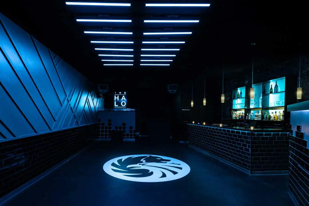 Interior-Fotografie eines schwach beleuchteter Dancefloor in einem Club mit blauer Beleuchtung, einem Drachenlogo auf dem schwarzen Boden, einer Bar mit beleuchteten Regalen auf der rechten Seite und geometrischen Wandgestaltungen auf der linken Seite. Auf der Rückseite leuchtet das Wort HALO auf.