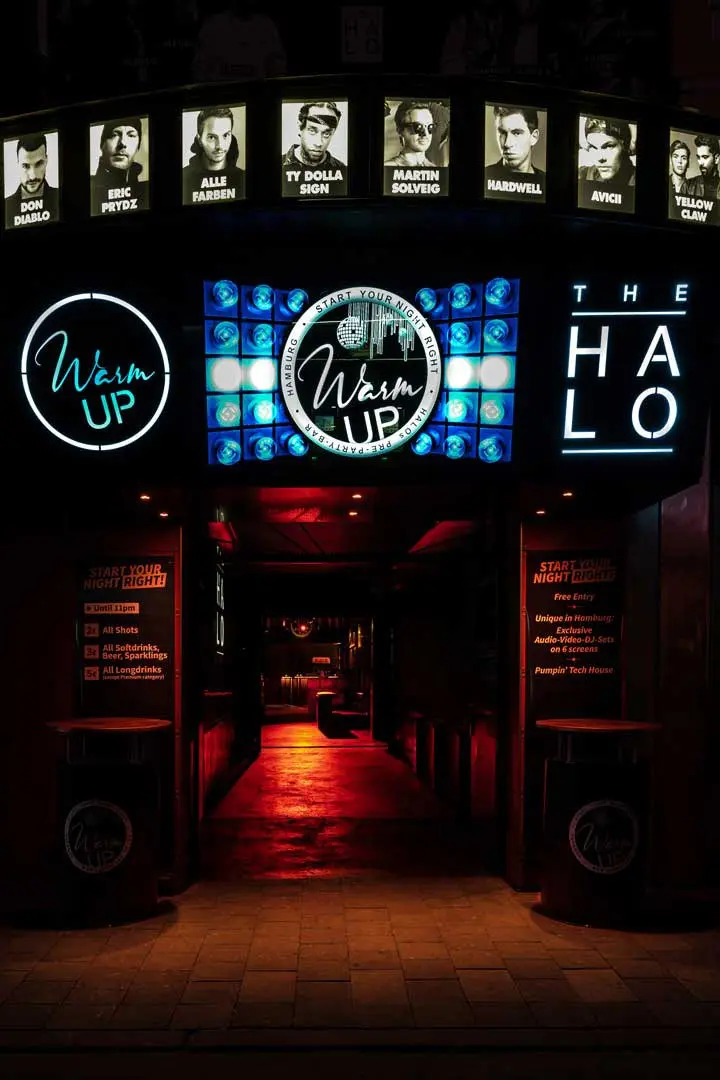 Eingang zu einem Nachtclub namens Warm Up und The Halo mit beleuchteten Schildern, Postern von DJs über dem Eingang und einem schwach beleuchteten Innenraum mit leuchtend rotem Boden.