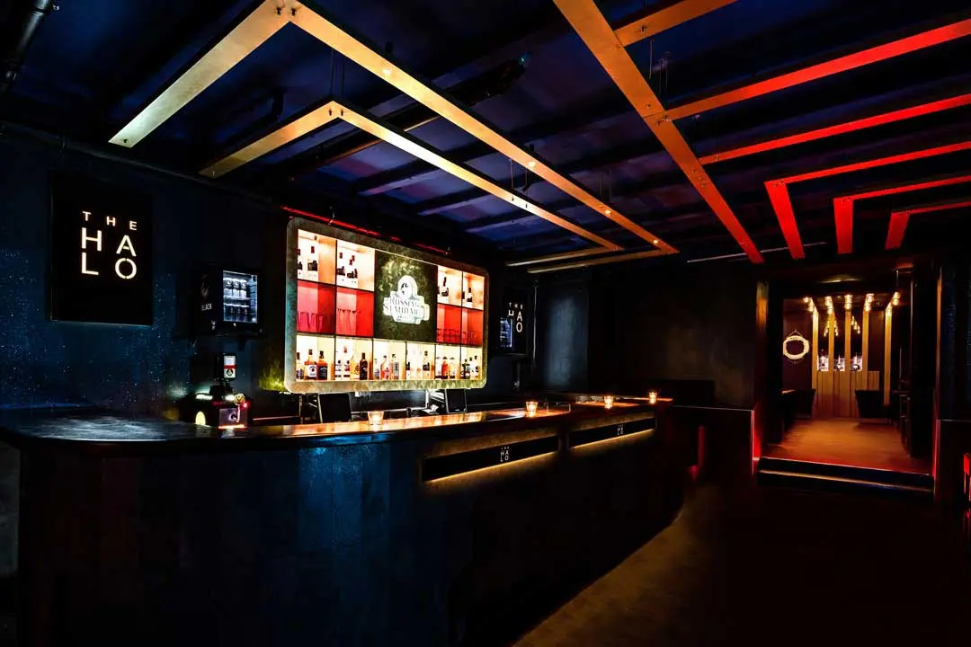 Eine moderne, schwach beleuchtete Bar in einem Club mit bunten, hinterleuchteten Regalen mit Flaschen hinter dem Tresen. Das dunkle Interieur wird durch eine warme Beleuchtung akzentuiert, mit einem Schild mit der Aufschrift The Halo an der Wand und geometrischen Deckenleuchten.
