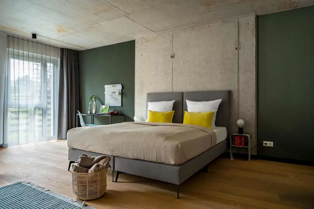 Modernes Schlafzimmer mit einer Akzentwand aus Beton, einem grauen Bett mit gelben Kissen, beiger Bettwäsche, einem Schreibtisch in der Nähe des Fensters, einem Weidenkorb mit Decken, einem Holzboden und durchsichtigen Vorhängen, die natürliches Licht hereinlassen.