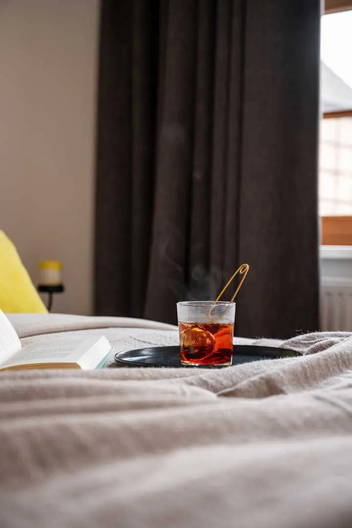Ein dampfendes Glas Tee mit einem Löffel und einer Zitronenscheibe steht auf einem Tablett auf einem gemütlichen Bett, neben einem aufgeschlagenen Buch. Sanftes Licht fällt durch ein Fenster mit dunklen Vorhängen im Hintergrund.